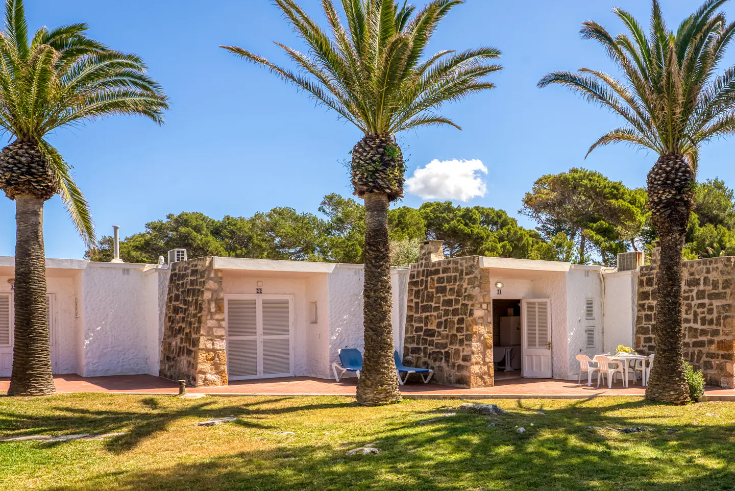 Exterior view of white bungalows with stone accents, palm trees, and lawn under a blue sky.