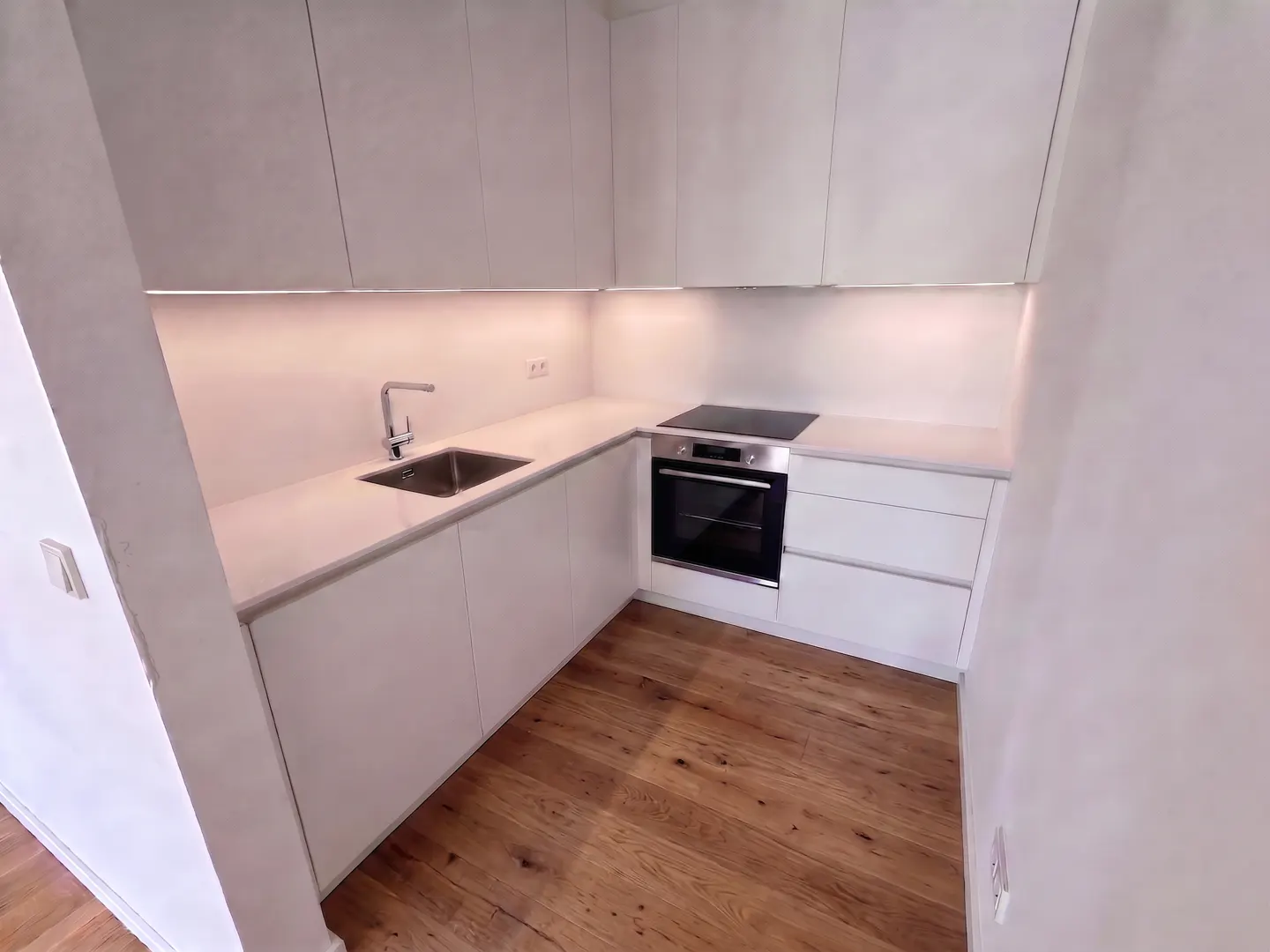 Bright kitchen with white cabinets, countertops, and walls. Stainless steel sink, faucet, oven, and cooktop. Wood floor.
