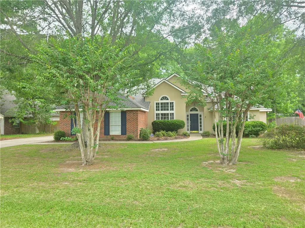 A single-story home with a brick and tan facade, dark blue shutters, and a green lawn. Trees frame the house.