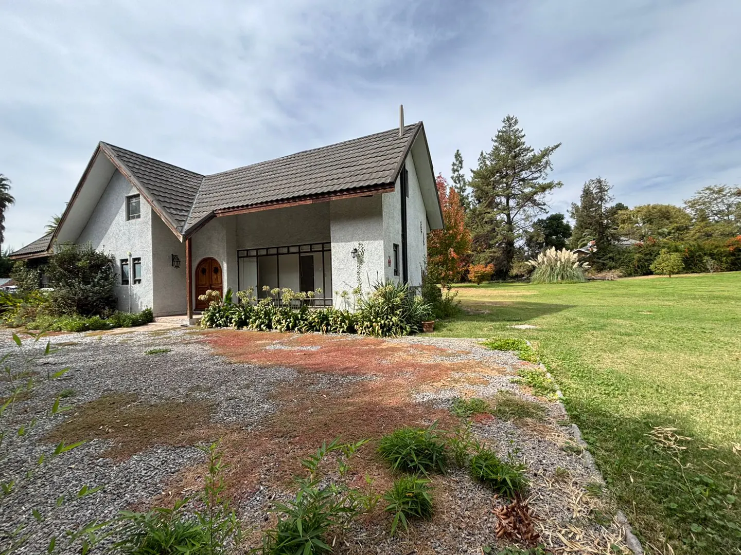 A white house with a gray roof sits on a gravel driveway next to a green lawn with trees.