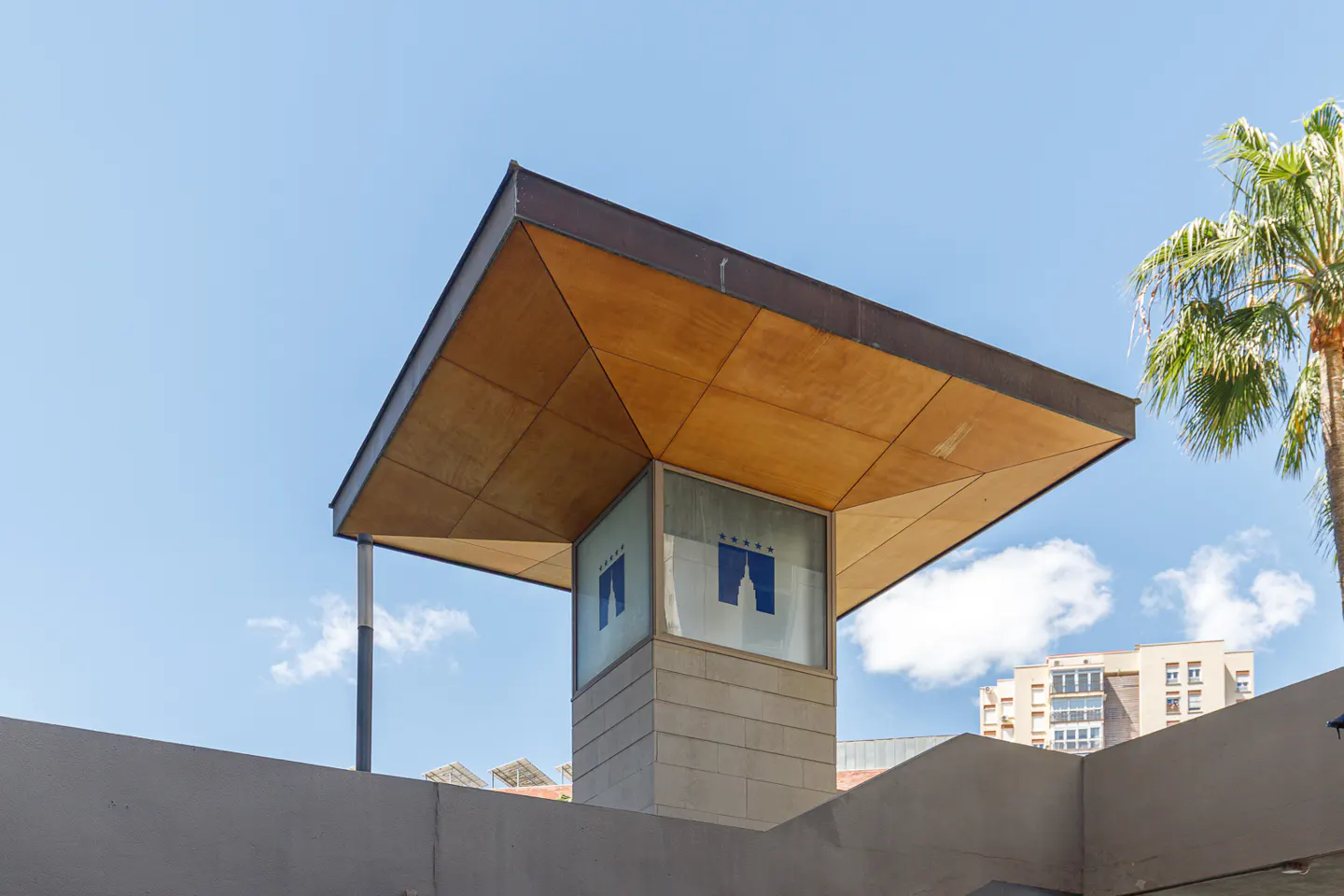 Modern building exterior with a brown roof, stone pillar with a blue logo, and a palm tree against a blue sky.