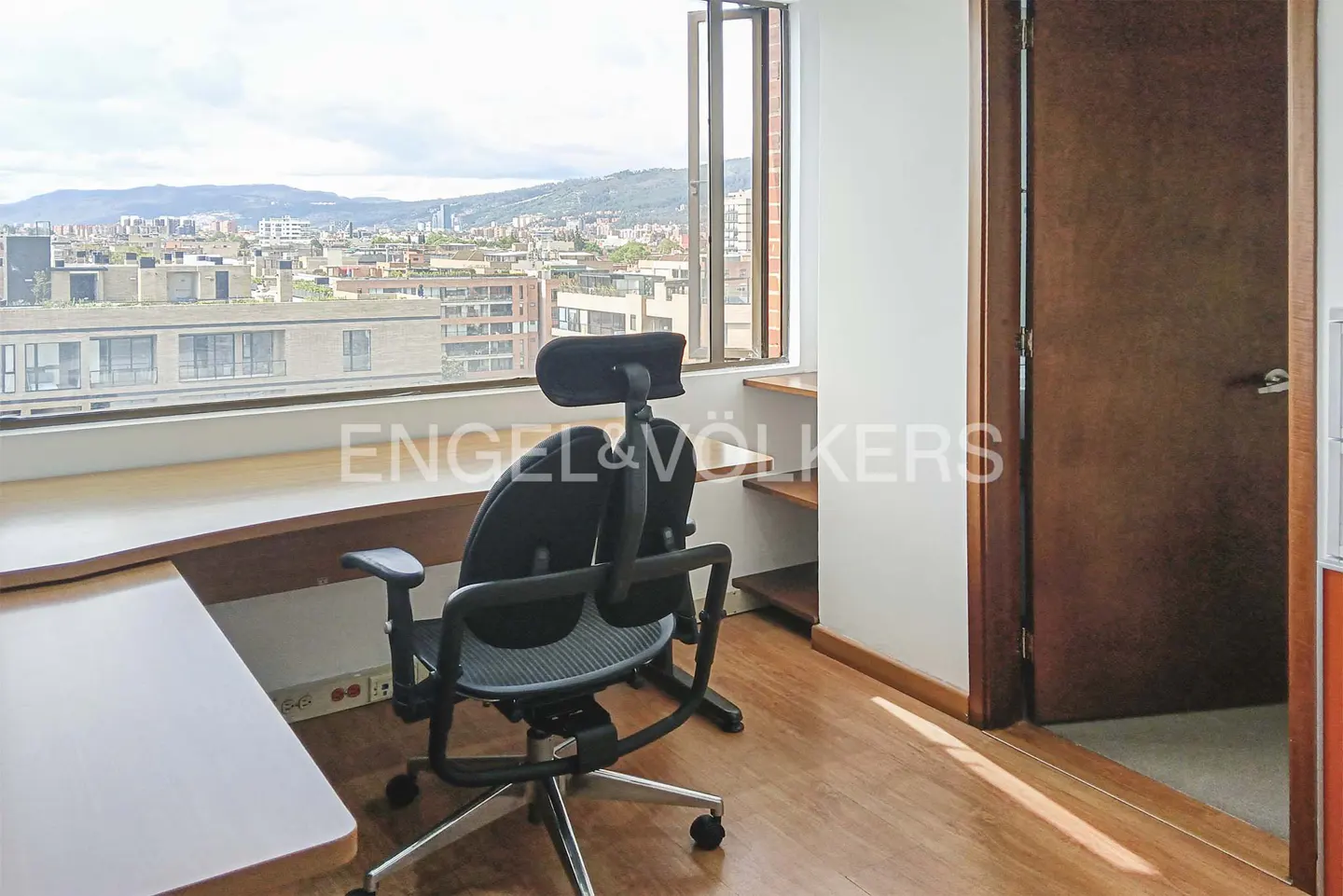 A bright office space with a black chair, wooden desk, and a large window overlooking a city. A brown door is on the right.