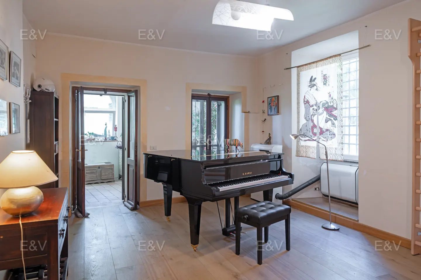 A bright room with a black grand piano and stool on a light wood floor. A window with a Japanese-style print is to the right.