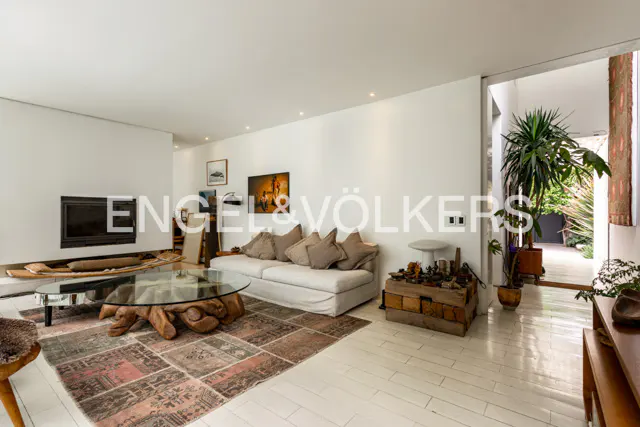 Bright living room with white walls, a fireplace, a white sofa, and a unique wooden coffee table on a patterned rug. A doorway leads to a lush outdoor area.