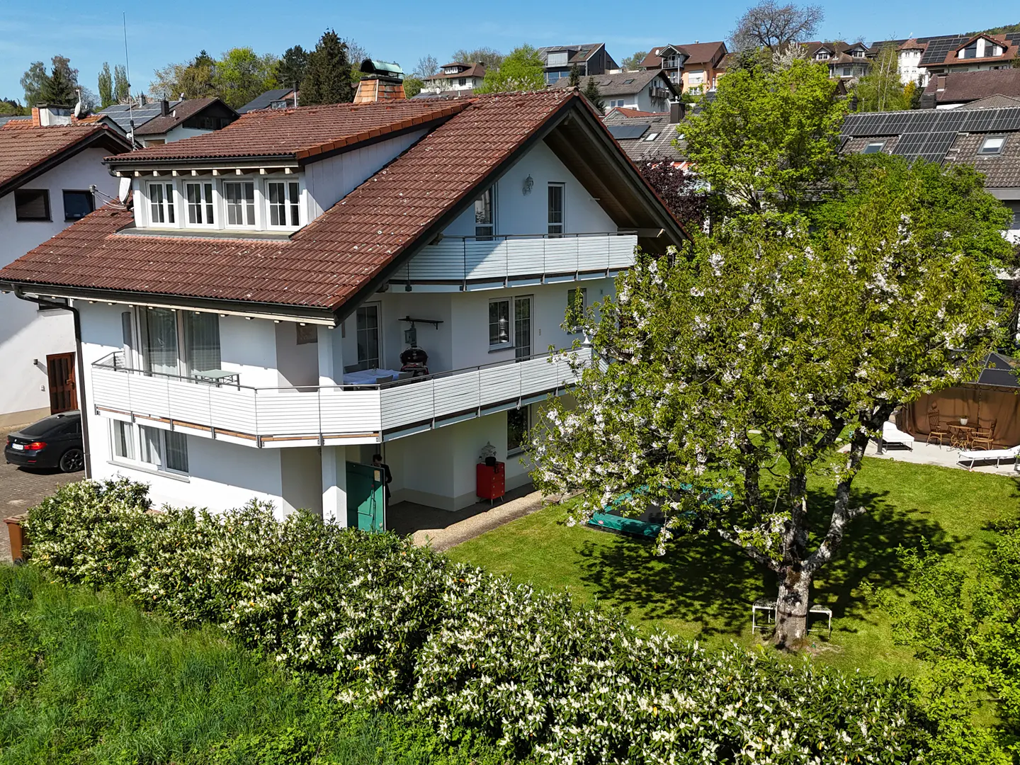 Two-story white house with a brown roof, balconies, green lawn, and a flowering tree.