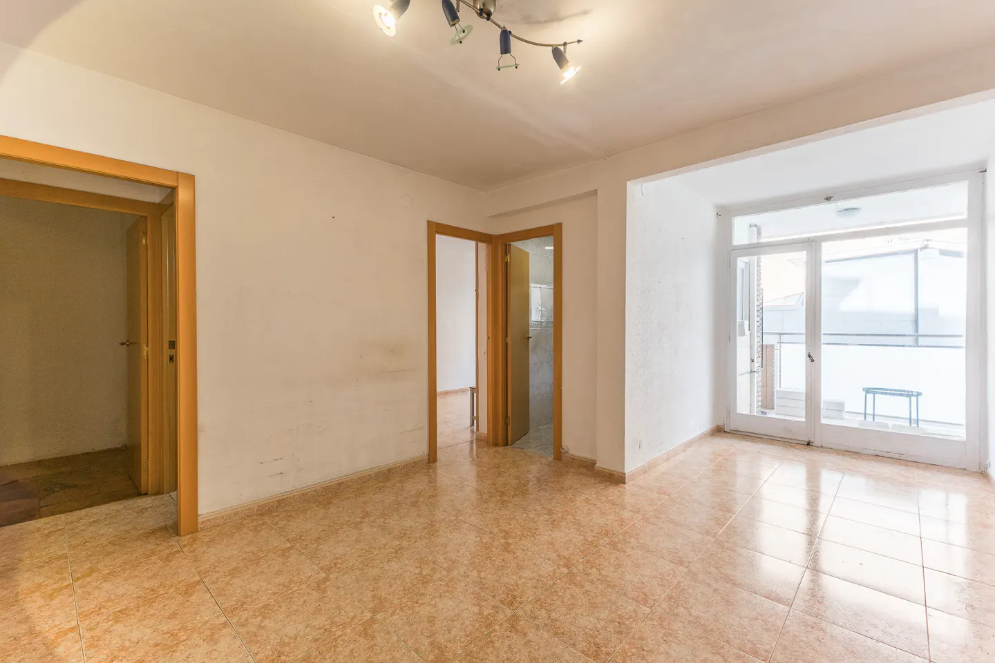 Empty room with tan tile floor, white walls, and wood trim. Doors lead to other rooms, and glass doors open to a balcony.