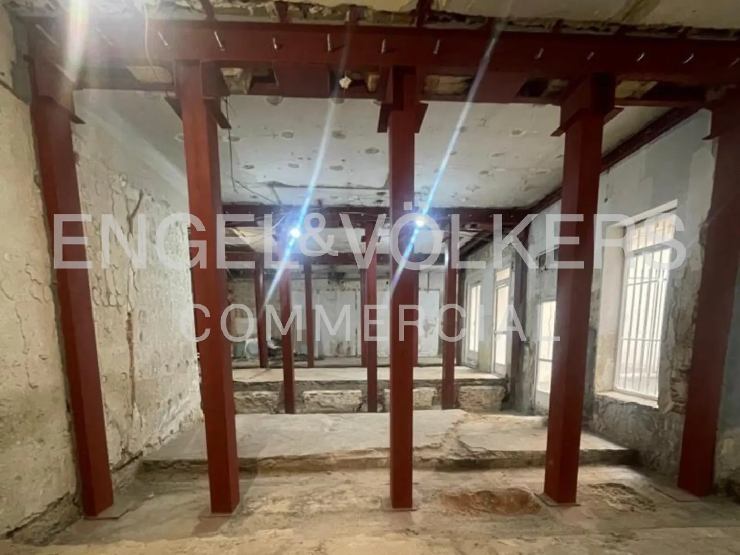 Interior view of a commercial space under renovation, featuring red support beams, exposed stone floors, and multiple windows. "Engel & Völkers Commercial" logo visible.