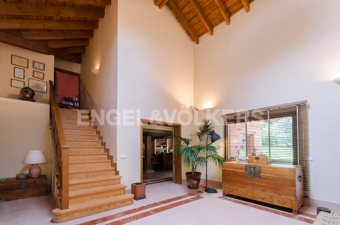 A bright foyer with a wooden staircase, a plant, and a wooden chest. The floor is tiled with red accents.