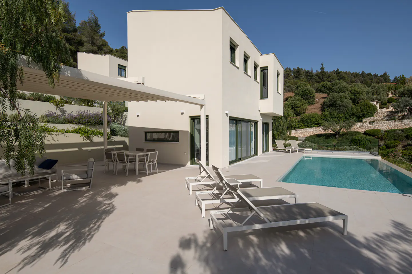 Modern white house with a pool, lounge chairs, and outdoor dining area. Trees and a blue sky are in the background.