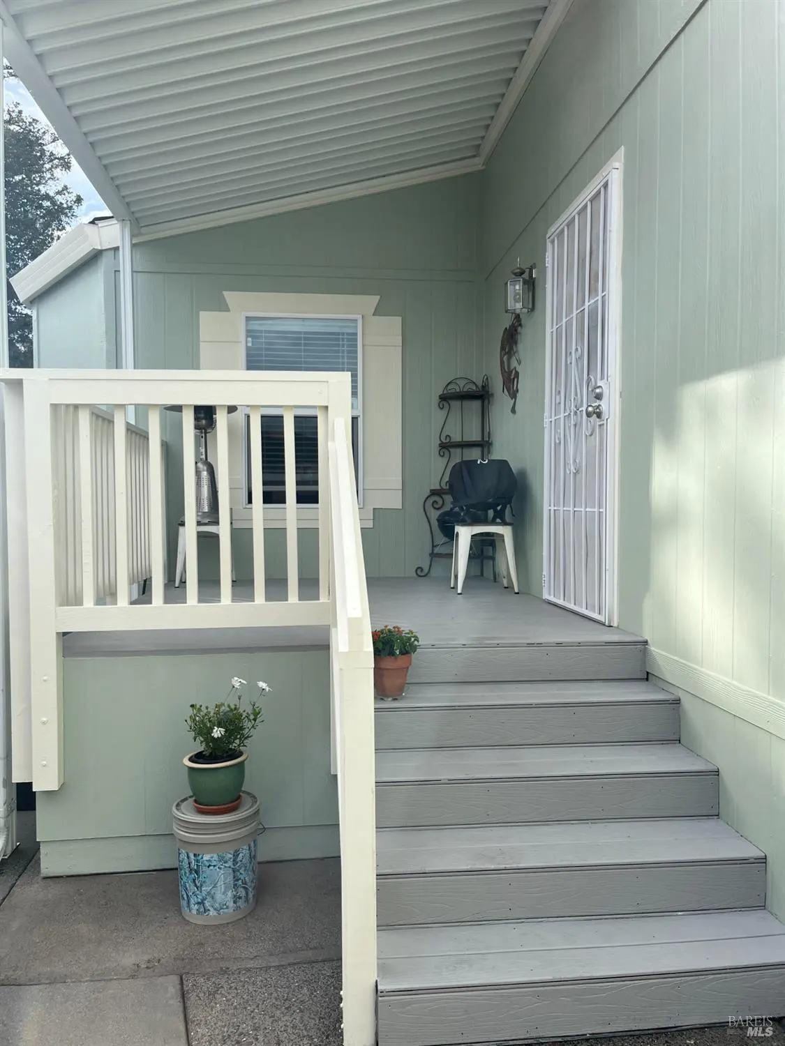 Exterior view of a light green home with white trim, steps leading to a covered porch with furniture and plants.