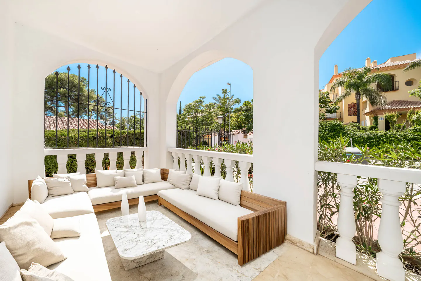 A bright, sunny patio with white walls, arched openings, and a wooden sofa with white cushions. A marble table sits in front, with a view of lush greenery and buildings.