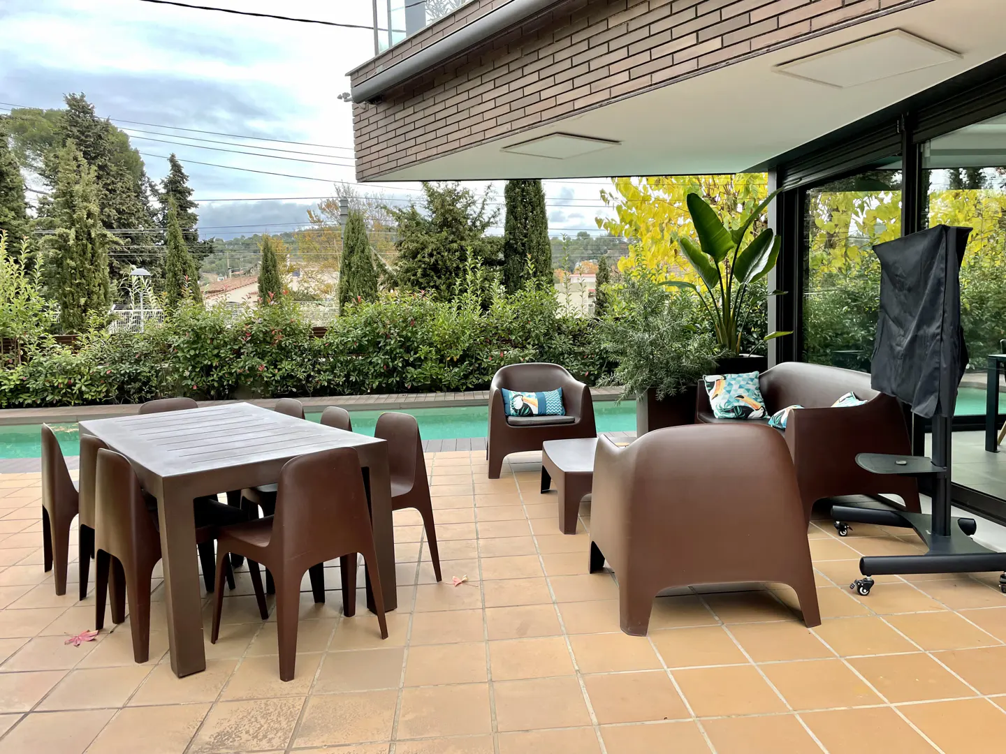 Outdoor patio with brown furniture, including a table with chairs and a lounge set, next to a pool and lush greenery.
