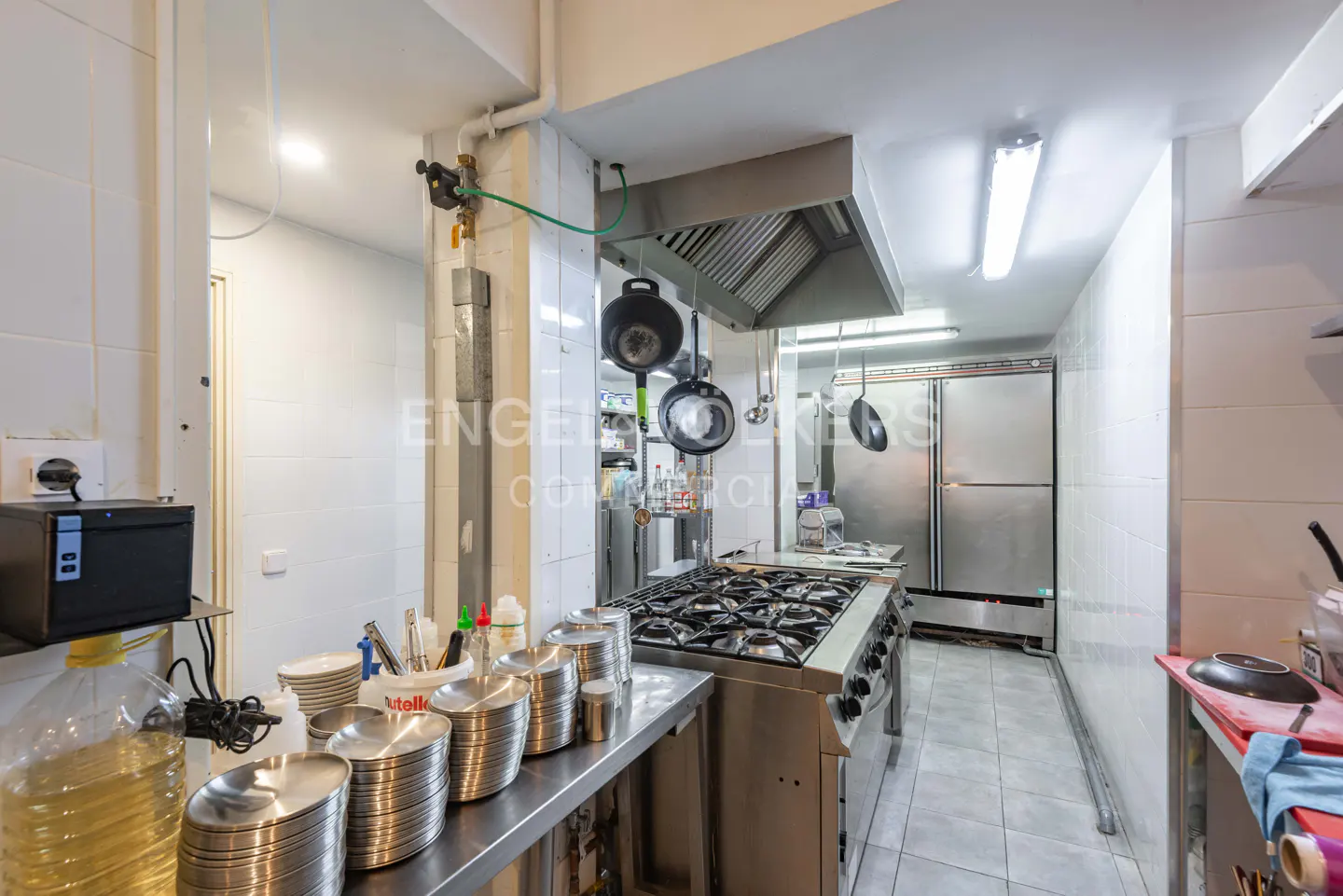 A commercial kitchen with stainless steel appliances, including a large stove, hood, and refrigerators. Utensils and containers are on the counter.