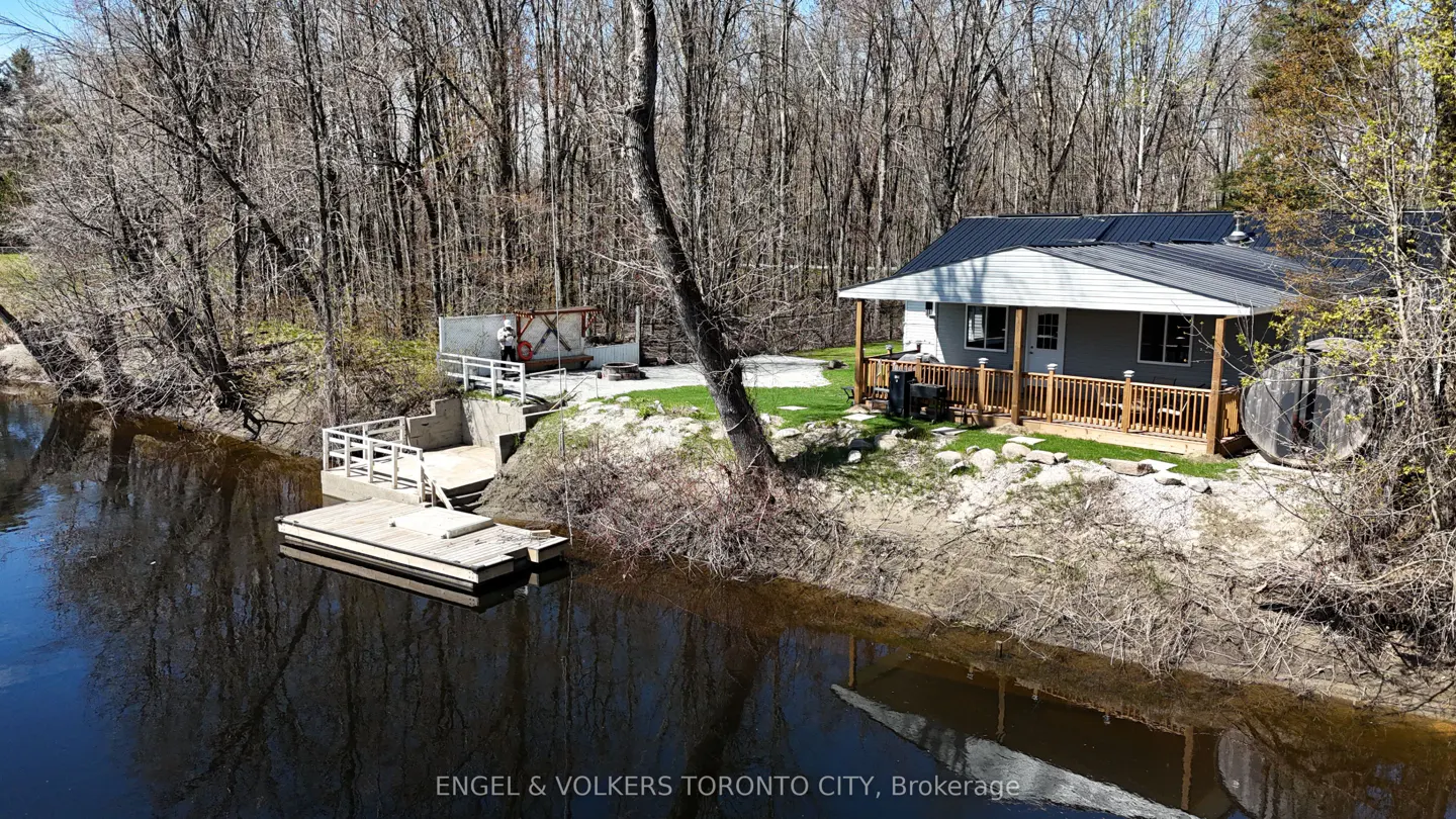 Aerial view of a gray house with a black roof and wooden porch, next to a river with a dock and concrete steps. Trees surround the property.
