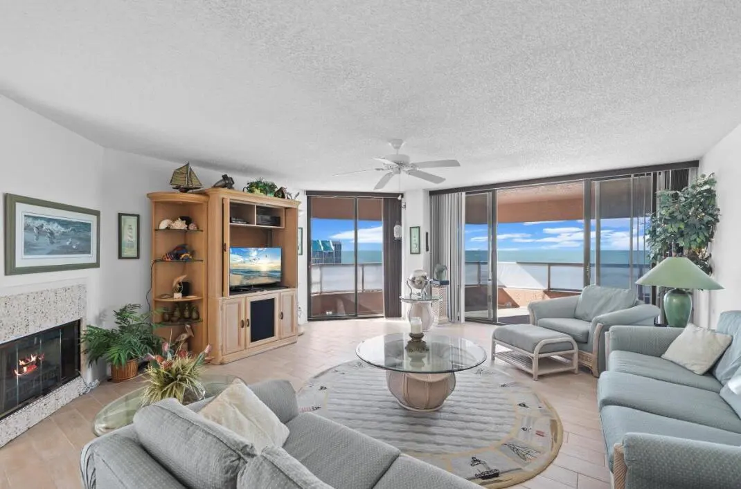 Living room with light blue sofas, a glass coffee table, and a view of the ocean through sliding glass doors. A fireplace and TV stand are also visible.