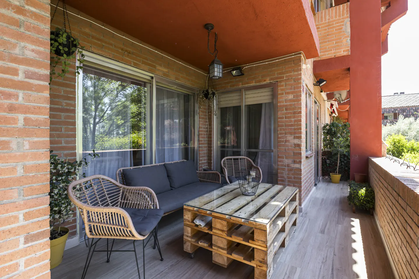 A cozy balcony with brick walls, a red ceiling, and outdoor furniture. A pallet table sits between a sofa and two wicker chairs.