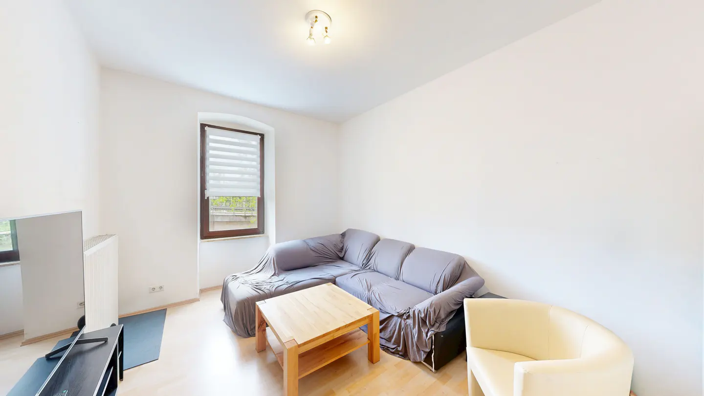 Bright living room with a gray sectional sofa, wooden coffee table, and a cream-colored armchair on a light wood floor. A window with blinds is on the left.