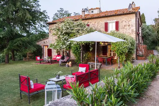 Stone house with red shutters and outdoor seating. Red chairs and tables are arranged on the lawn. A person sits in a hanging chair.