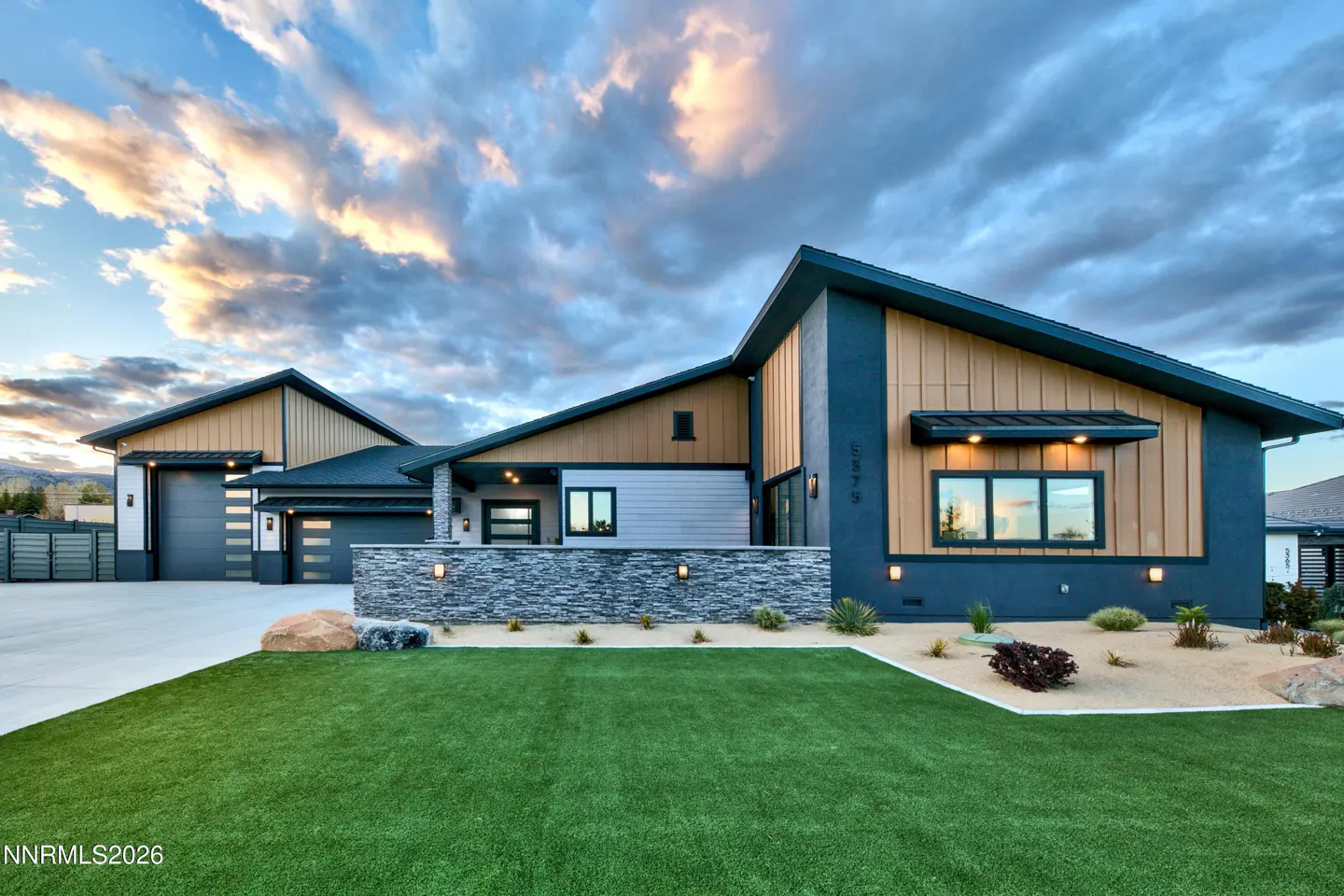 Modern home exterior with blue and tan siding, stone accents, green lawn, and cloudy sky at dusk.