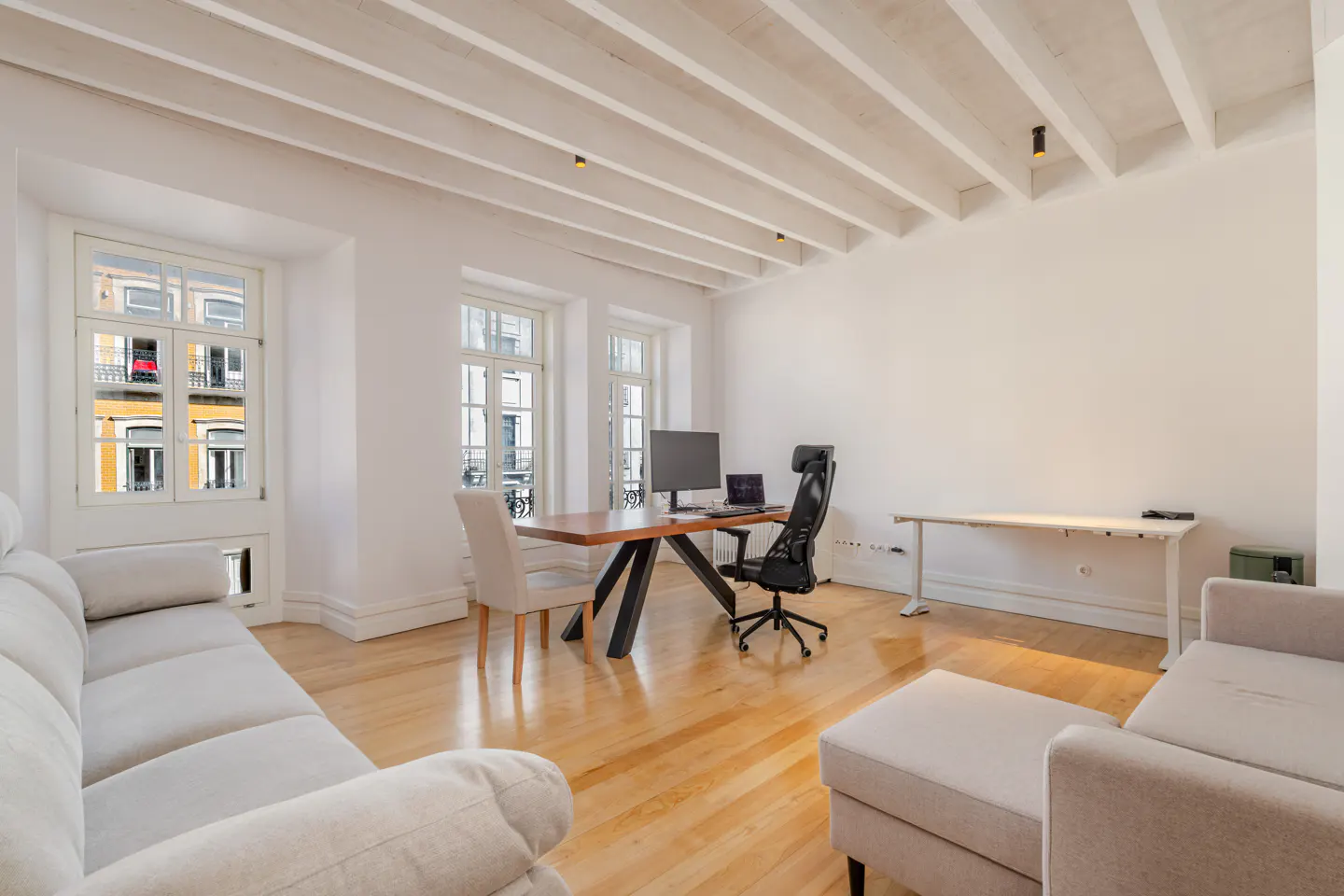 Bright, modern living room with white walls, wood floors, and exposed ceiling beams. A desk with computer and sofa create a comfortable workspace.