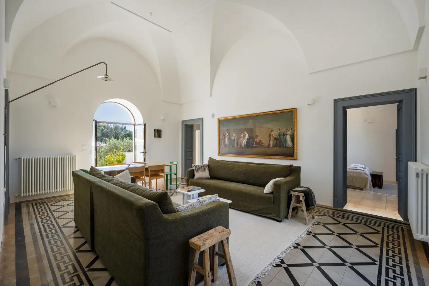 Living room with vaulted ceilings, olive green sofas, patterned tile floors, and a large painting on a white wall.