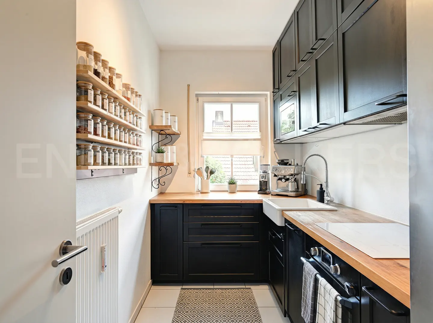 A bright kitchen with black cabinets, wood countertops, and open spice rack shelves. A window provides natural light.