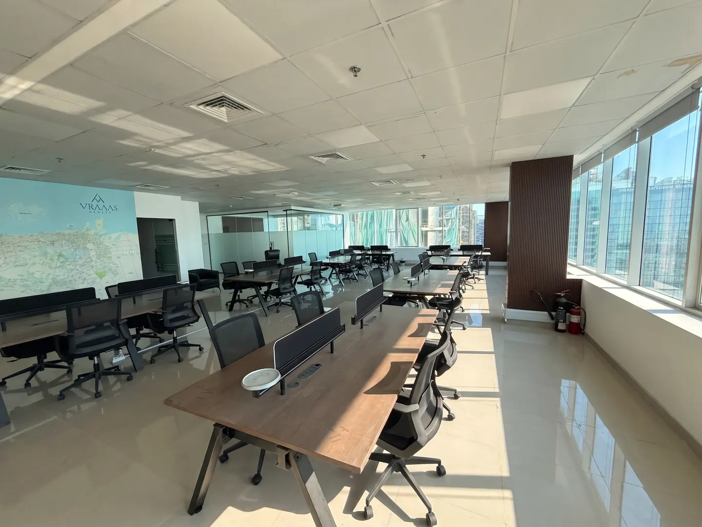 Bright, modern office space with long desks, black chairs, and large windows overlooking a city skyline. A map mural is on the wall.