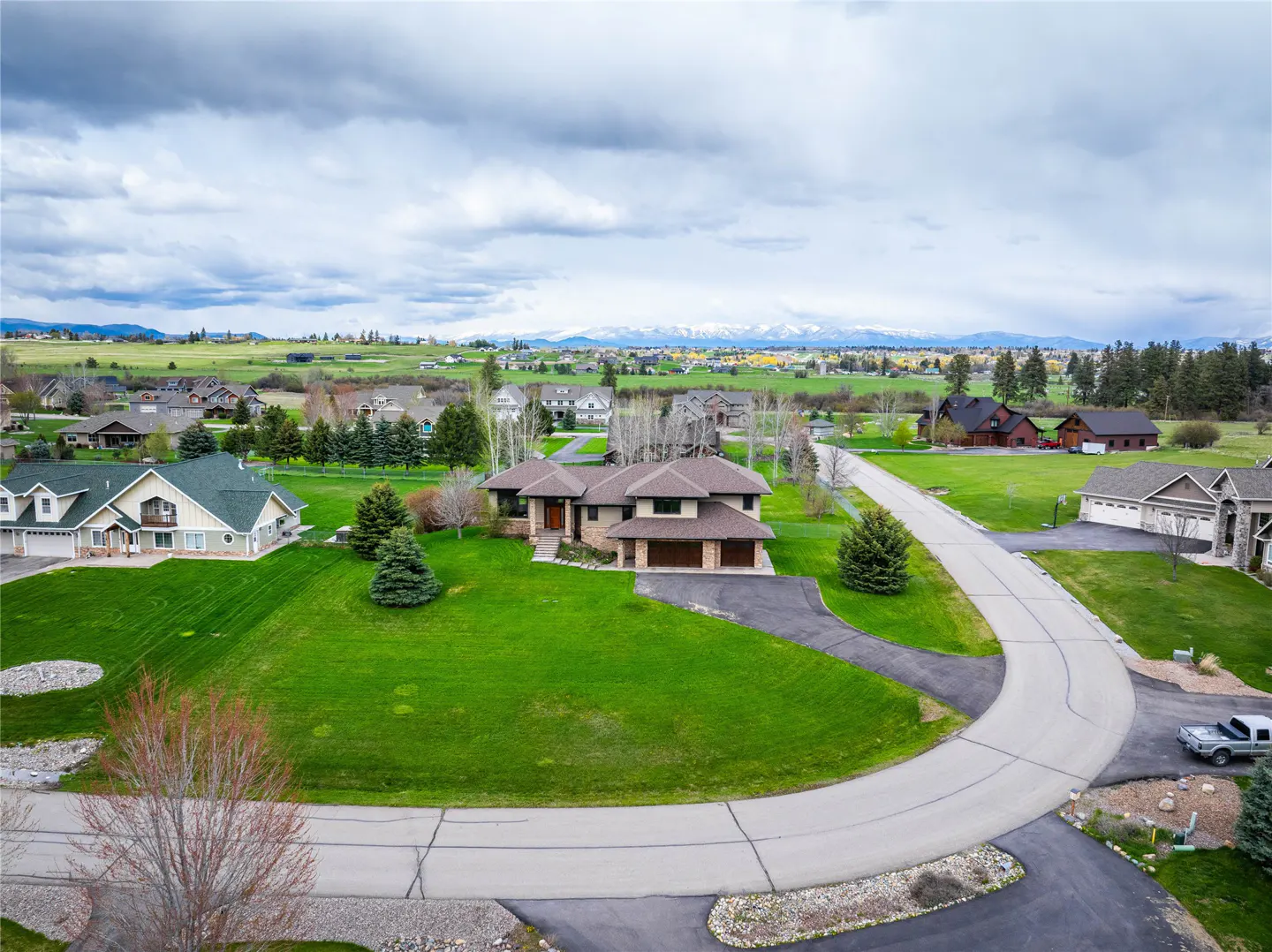 Aerial view of a tan house with a brown roof, green lawn, and a curved driveway in a rural neighborhood. Snow-capped mountains are in the distance.