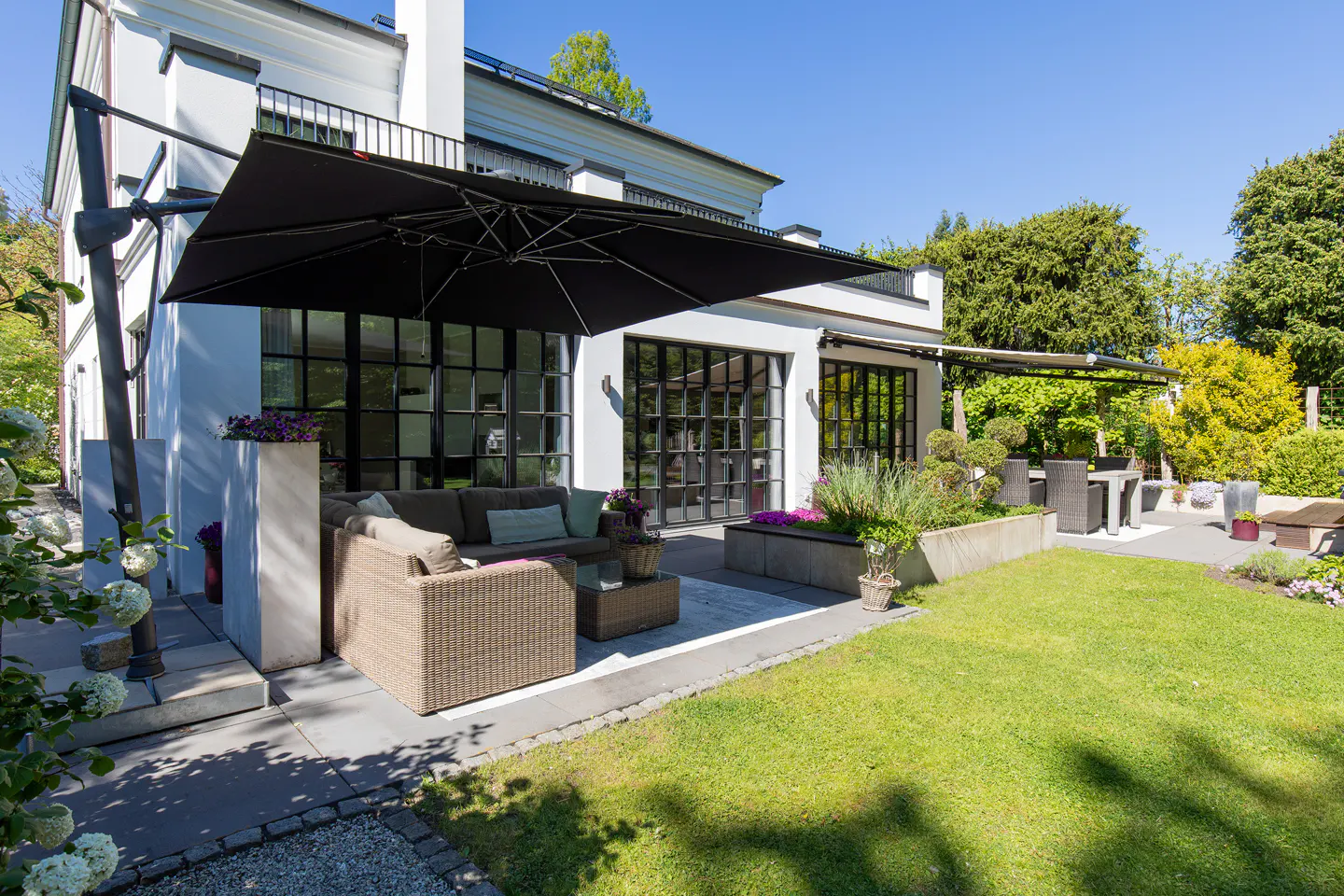 A modern white house with black framed windows, a patio with wicker furniture, and a large black umbrella. Green lawn and trees surround the house.