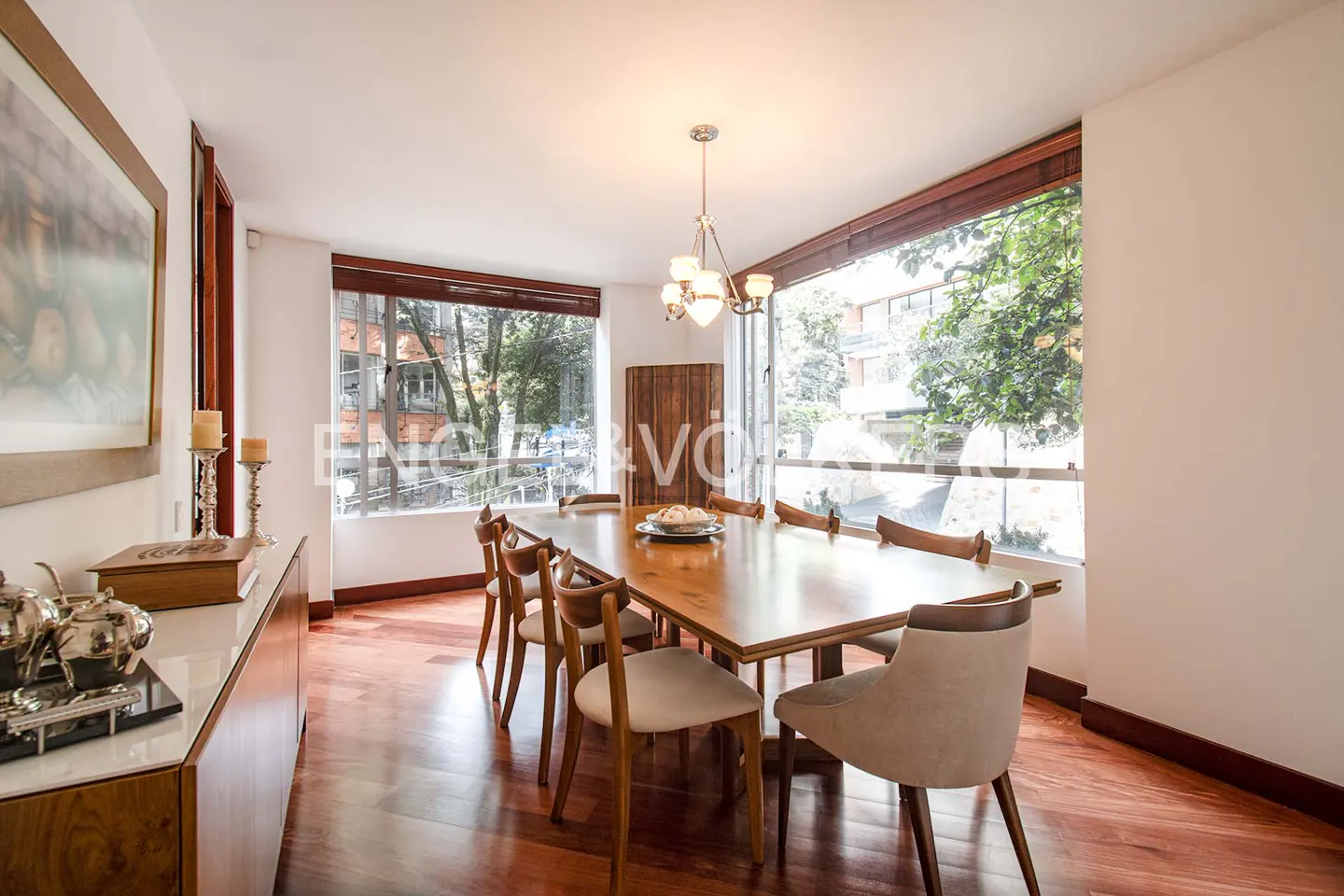 Bright dining room with wood floors, a long table with chairs, and large windows overlooking trees. A chandelier hangs above the table.