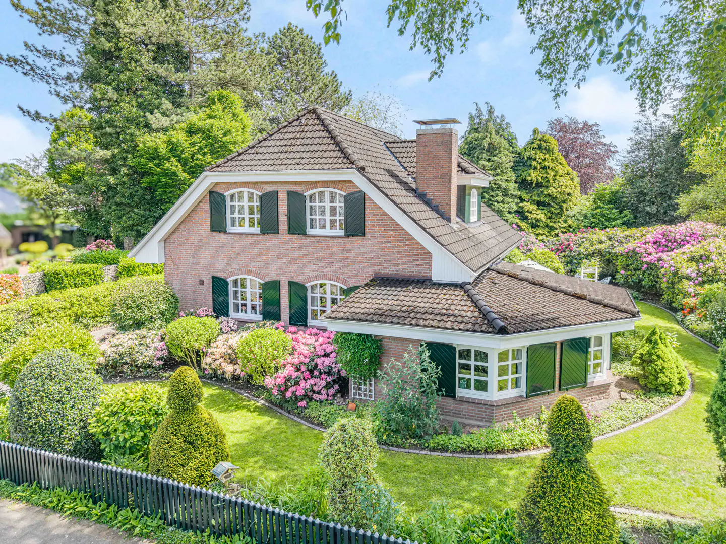 Brick house with dark green shutters and a brown tiled roof, surrounded by lush green landscaping and colorful flowers.