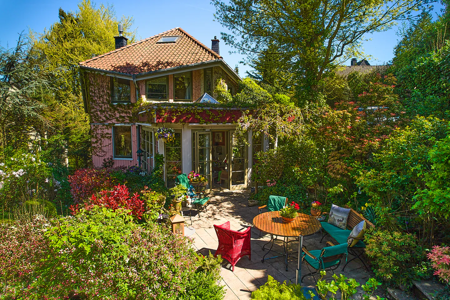 A pink house with a red tile roof is surrounded by lush green trees and bushes. A patio with outdoor furniture sits in front of the house.