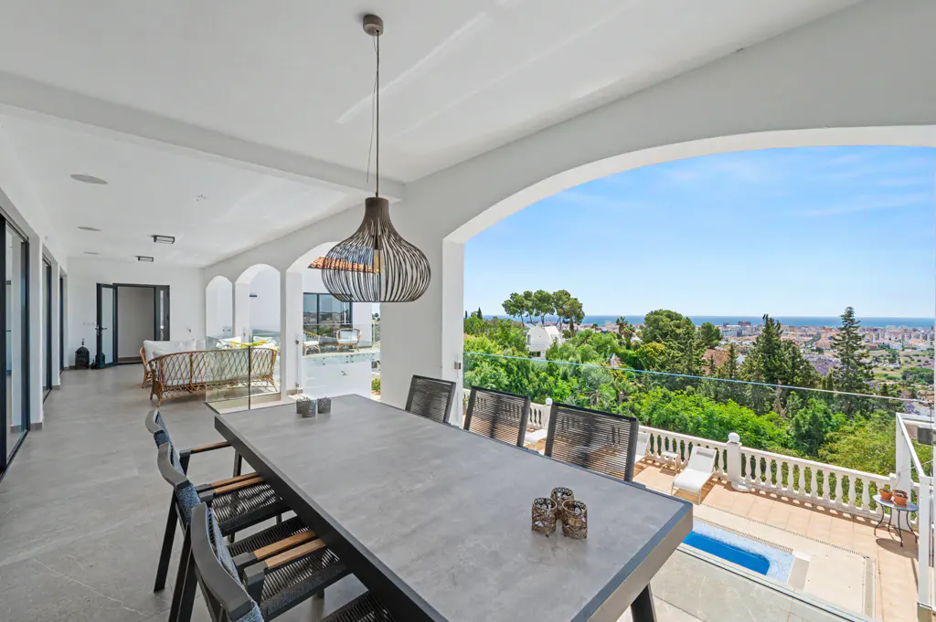 Covered patio with a long gray table, black chairs, and a modern light fixture. A view of trees, city, and ocean is in the background.