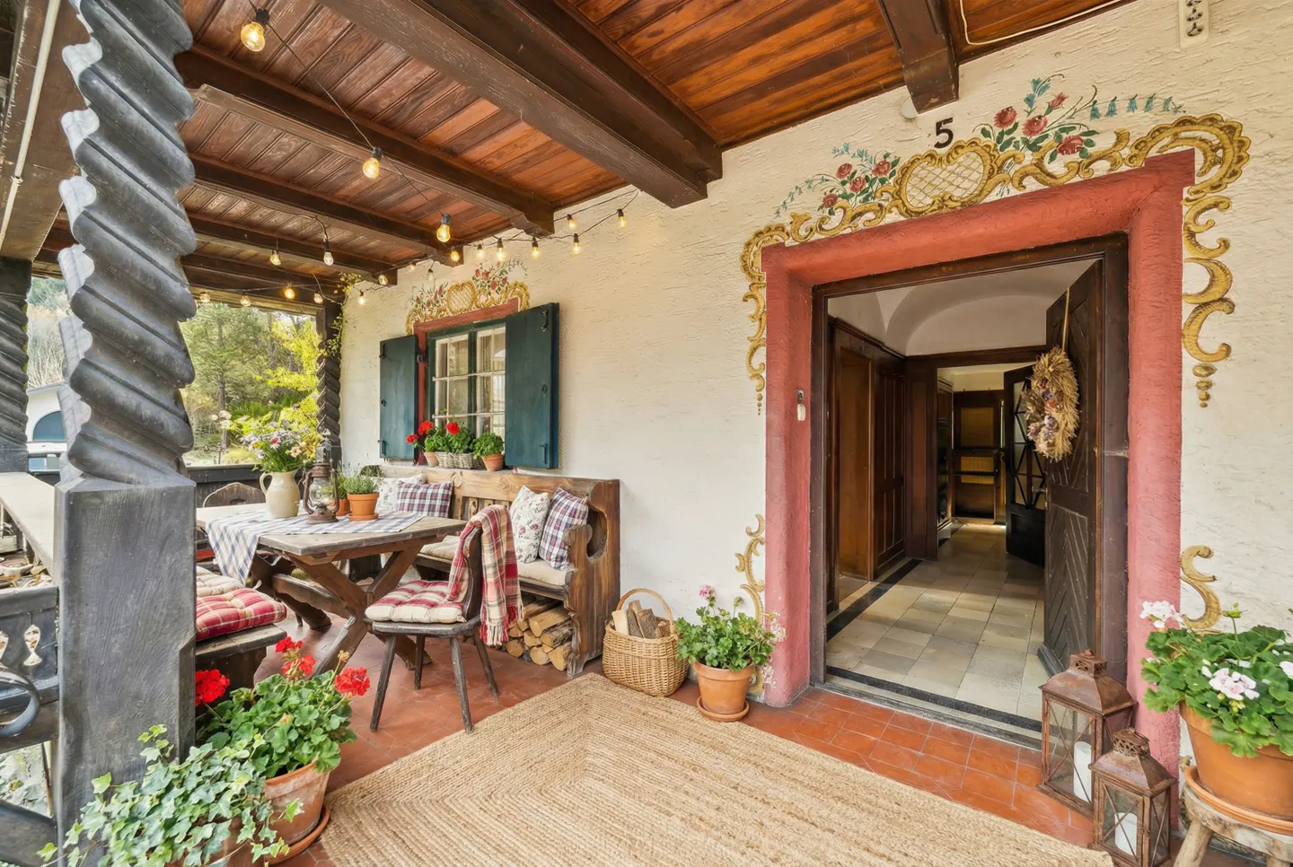 Covered porch with a table, chairs, and bench. String lights hang from the wooden ceiling. The door is decorated with floral designs.