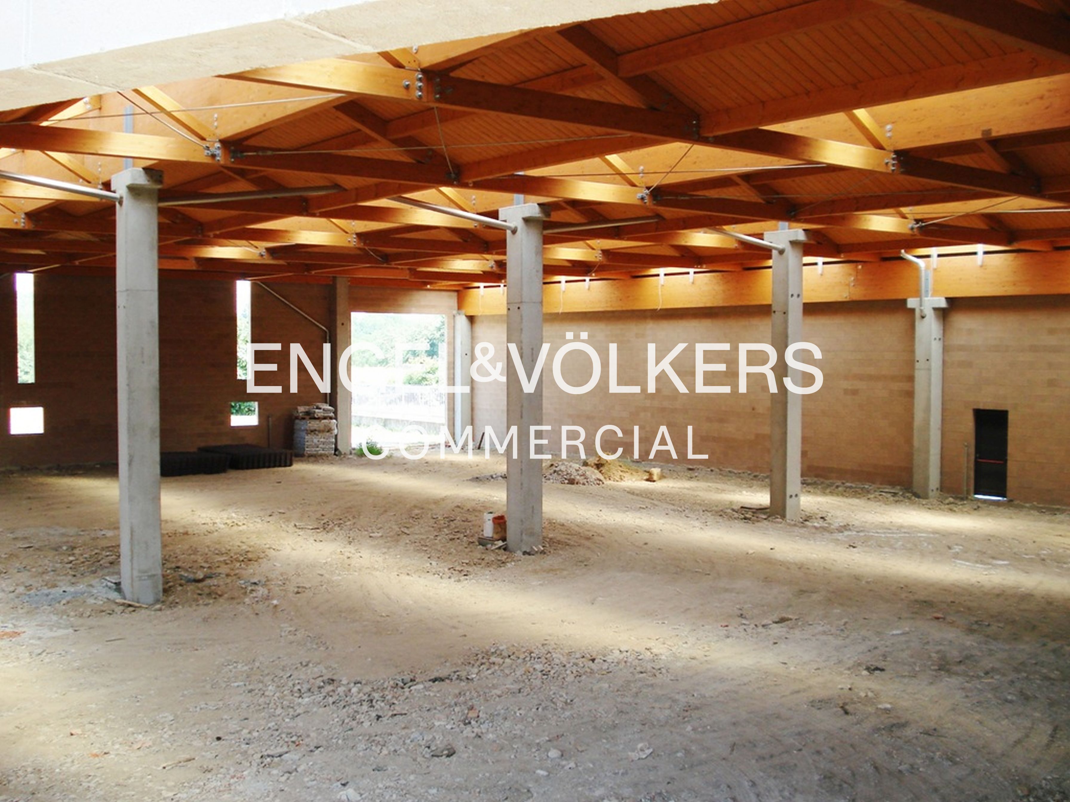 Large, unfinished commercial space with concrete pillars, tan brick walls, and an exposed wooden truss ceiling. [AI-generated text]