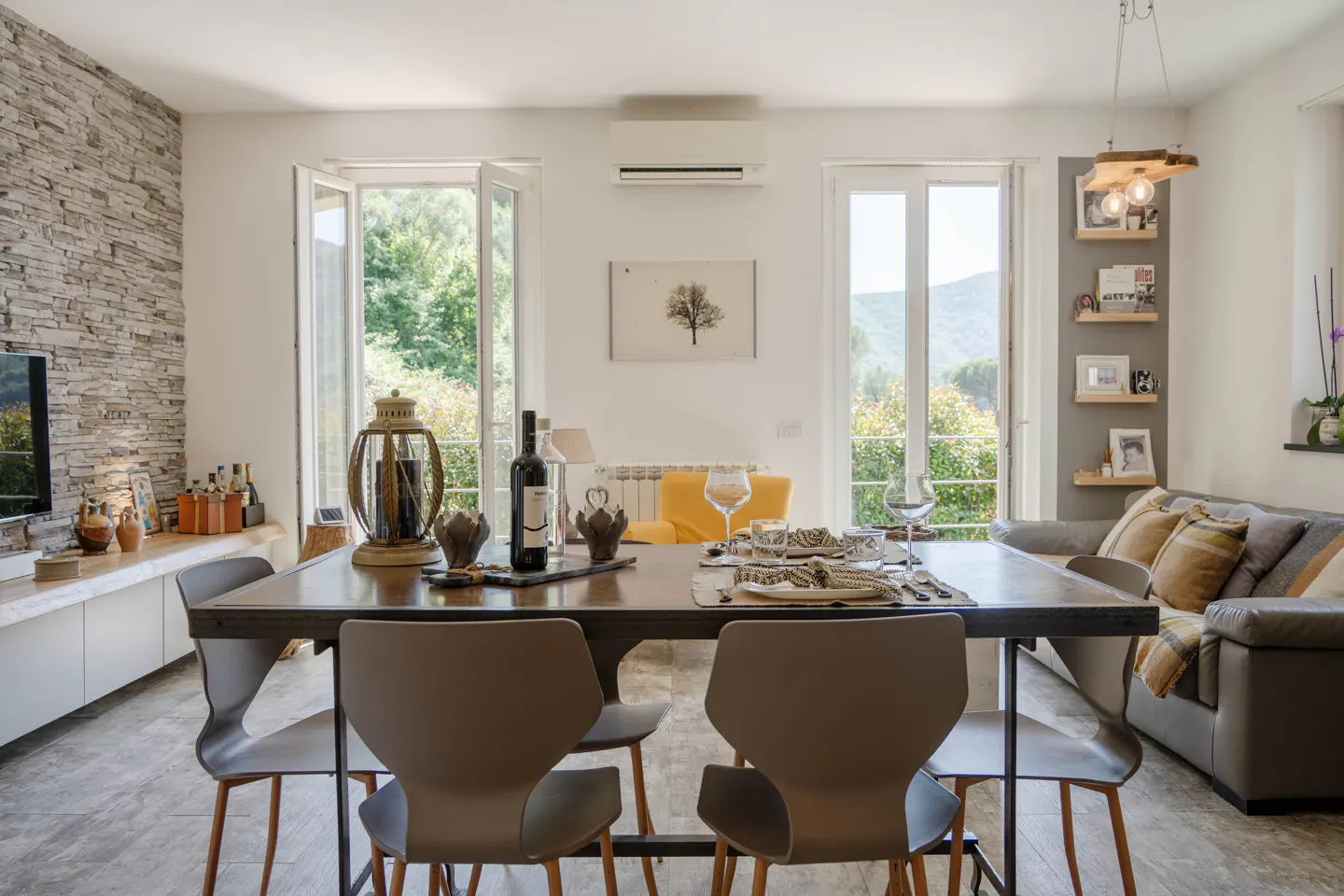 Bright dining area with a long wooden table set for a meal, surrounded by gray chairs. Windows overlook a lush green landscape.