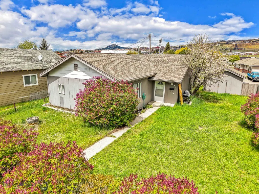 A single-story gray house with a green lawn and flowering bushes under a blue sky with white clouds.