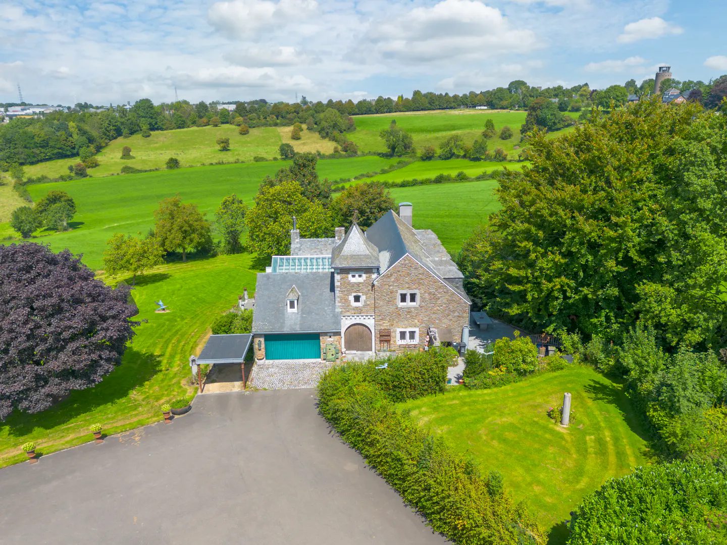 Aerial view of a stone house with a turret, green garage door, and a slate roof, surrounded by green lawns and trees. Rolling green hills in the background.