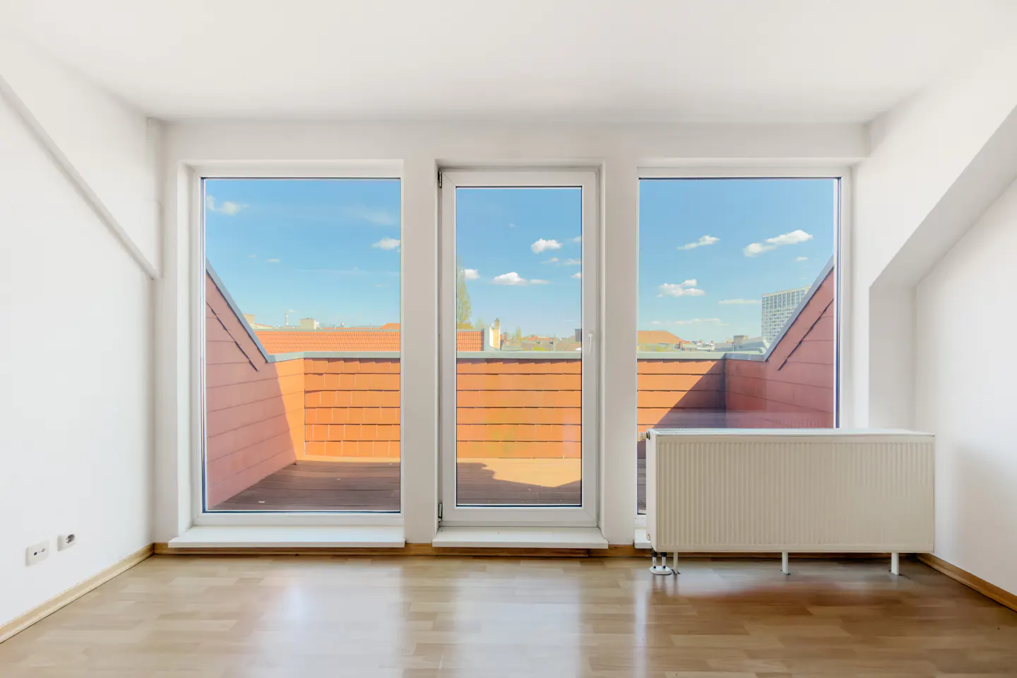 Bright, empty room with wood floor, white walls, and three windows showing a rooftop view under a blue sky. A white radiator sits by the right window.