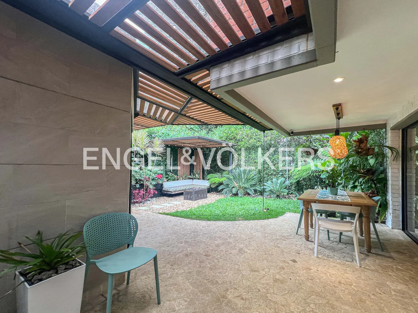 Outdoor patio with a wooden pergola, dining table, and a blue chair. A garden with a gazebo is visible in the background.