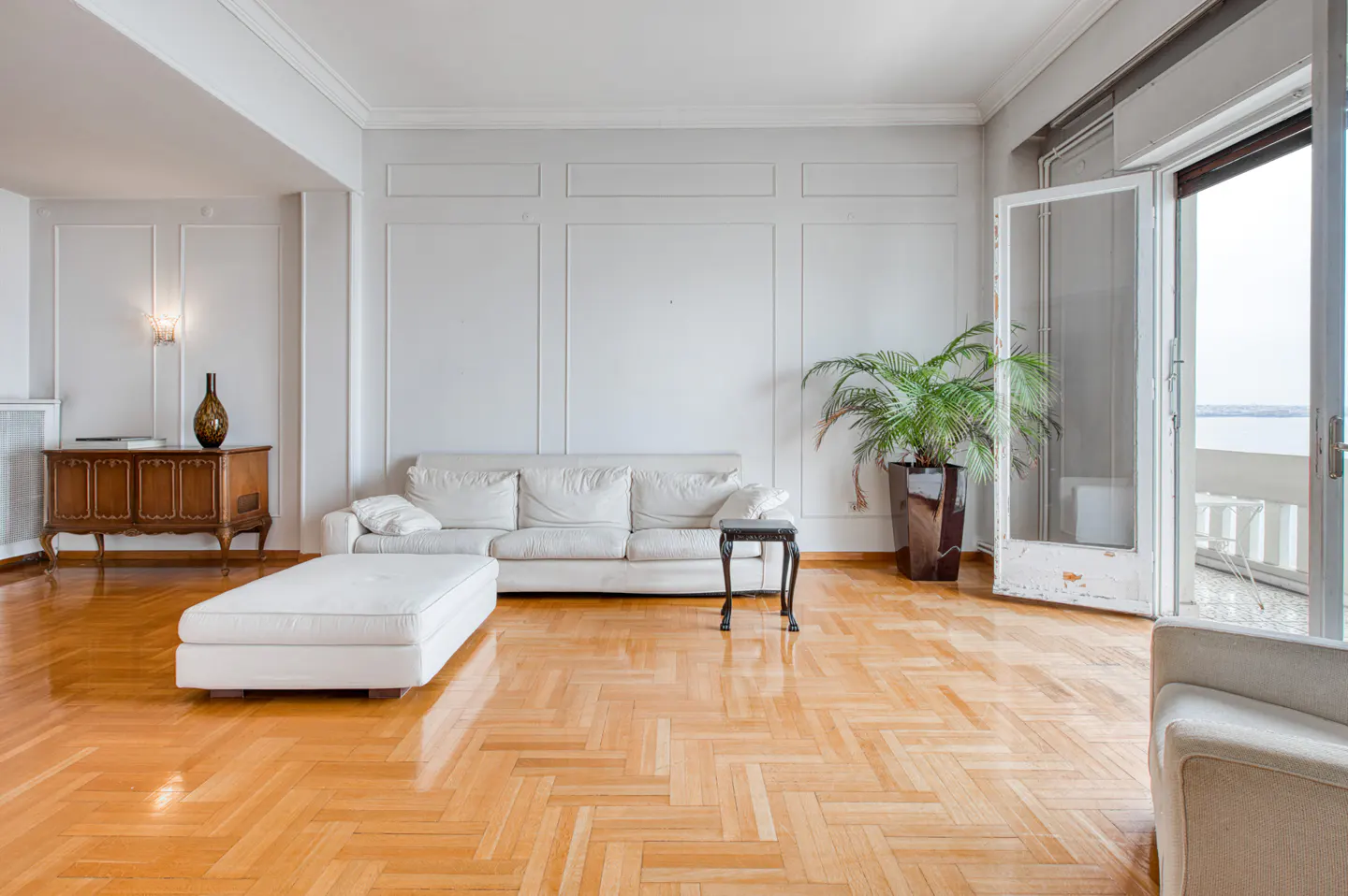 Bright living room with herringbone wood floors, white sofa, ottoman, and a potted palm near open balcony doors.