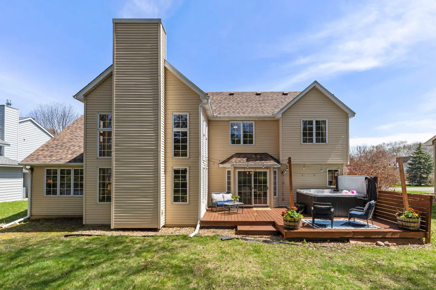Backyard view of a two-story beige house with a deck, hot tub, and green lawn.