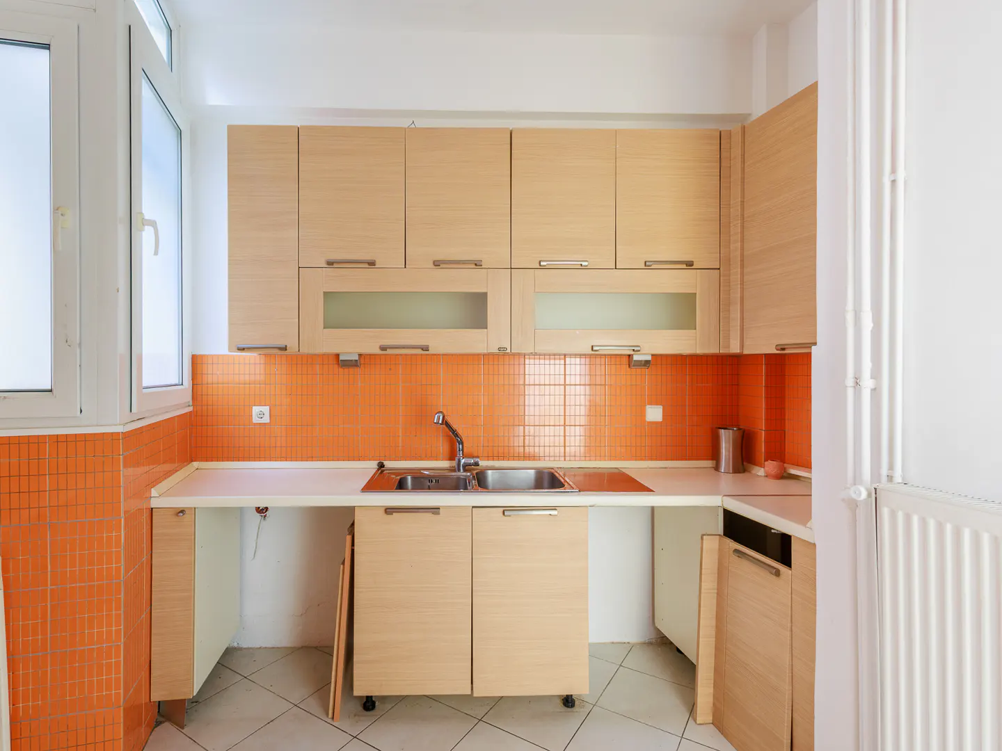 Bright kitchen with light wood cabinets, orange tile backsplash, and a stainless steel sink. A white window is on the left.