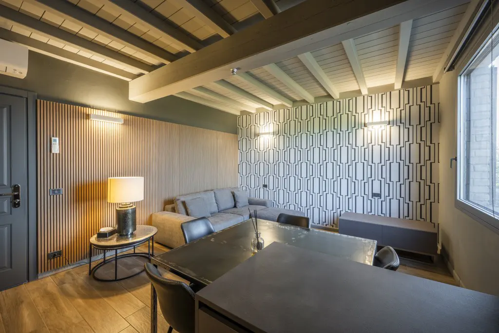 Living room with gray sofa, black table and chairs, wood floors, and patterned wallpaper. Beamed ceiling and large window.