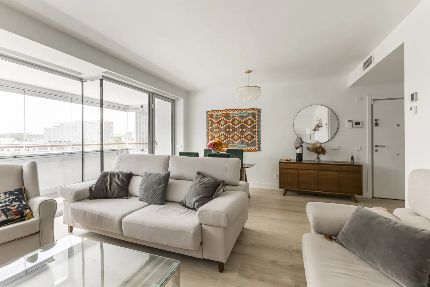 Bright living room with beige sofas, gray pillows, and a glass coffee table. A colorful rug hangs above a dining table with green chairs. A wooden cabinet sits near a round mirror.