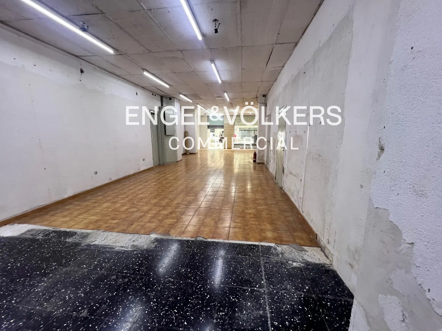 Empty commercial space with brown tile floor, white walls, and fluorescent lighting. Engel & Volkers logo is superimposed on the image.