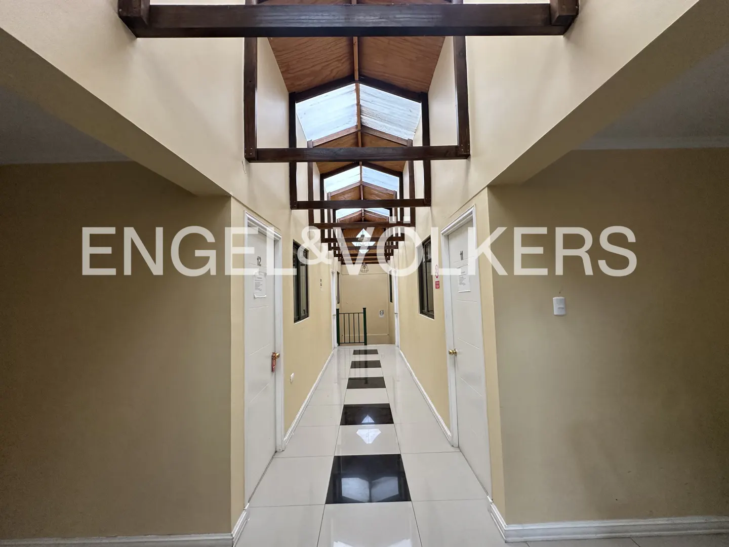 Hallway with beige walls, white doors, and a black and white tiled floor. A wooden beam structure supports a skylight ceiling. Engel & Volkers logo is superimposed.