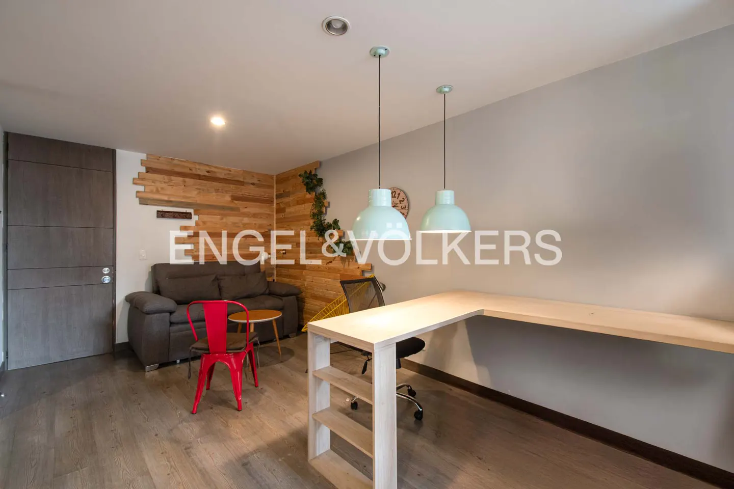 Living room with gray sofa, red chair, and wooden desk under two light blue pendant lamps. Wood paneled wall and gray door.