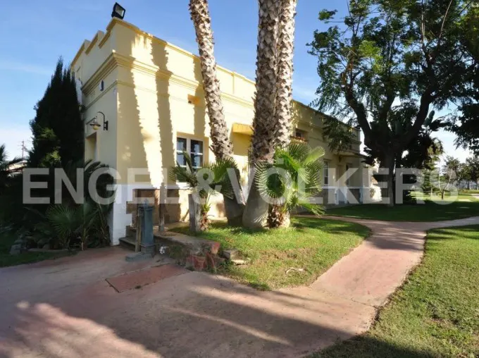 Exterior view of a yellow, two-story house with palm trees and a red brick walkway. The sky is blue and sunny.
