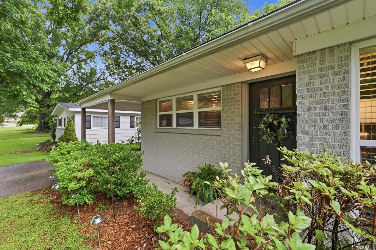 Exterior view of a one-story, light gray brick house with a dark front door and a green wreath. Green bushes in the front yard.